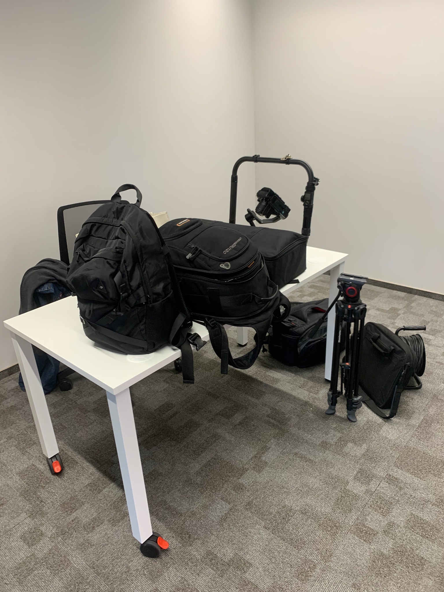 Sprzęt wideo na białym biurku: plecaki, statyw z kamerą, stabilizator. Przygotowania do nagrania w studio z szarą wykładziną.