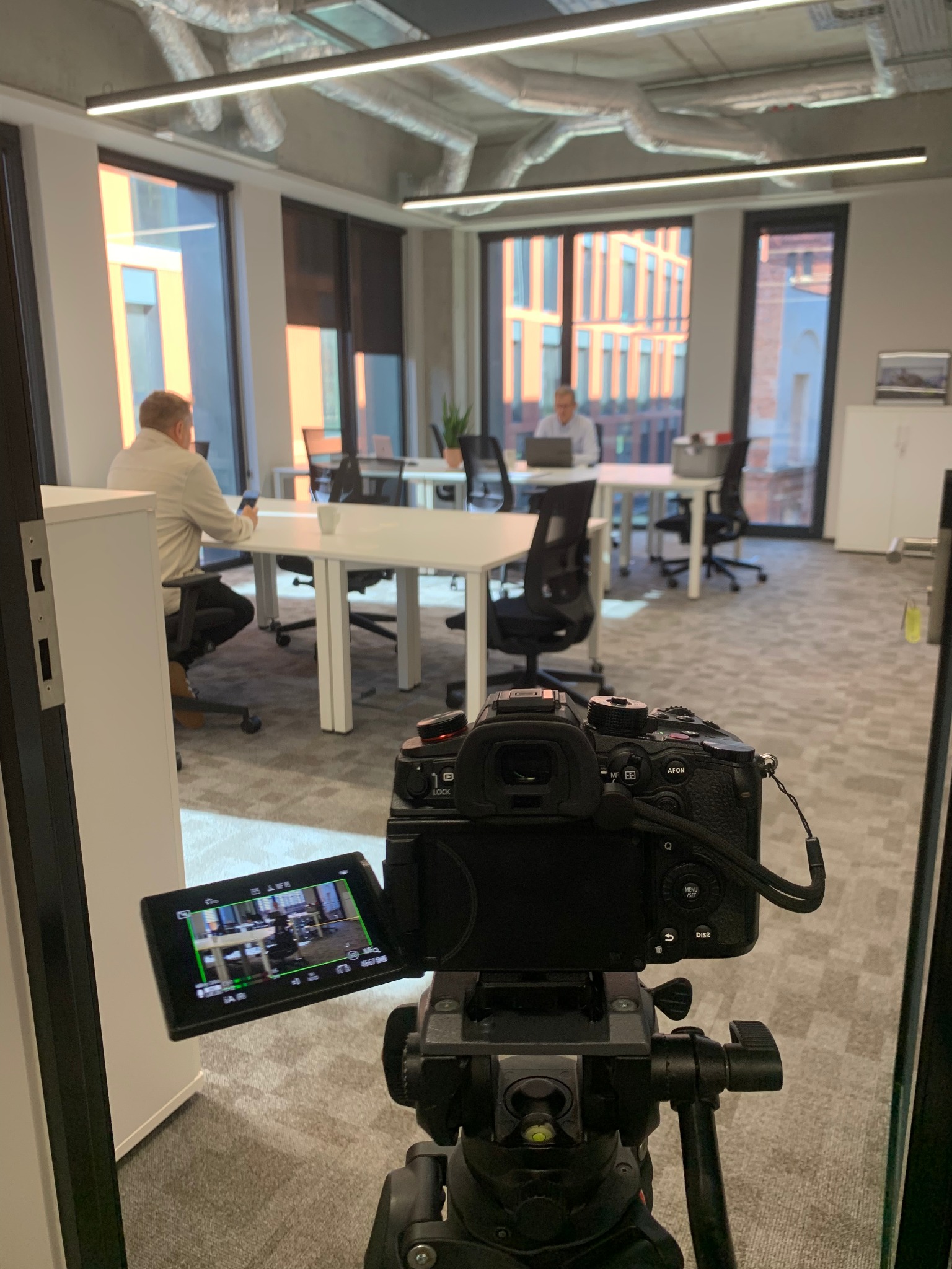 Kamera na statywie nagrywa wnętrze biura z pracującymi osobami. Widok zza kamery, z ekranem podglądowym. Nowoczesne biuro z dużymi oknami i białymi biurkami.