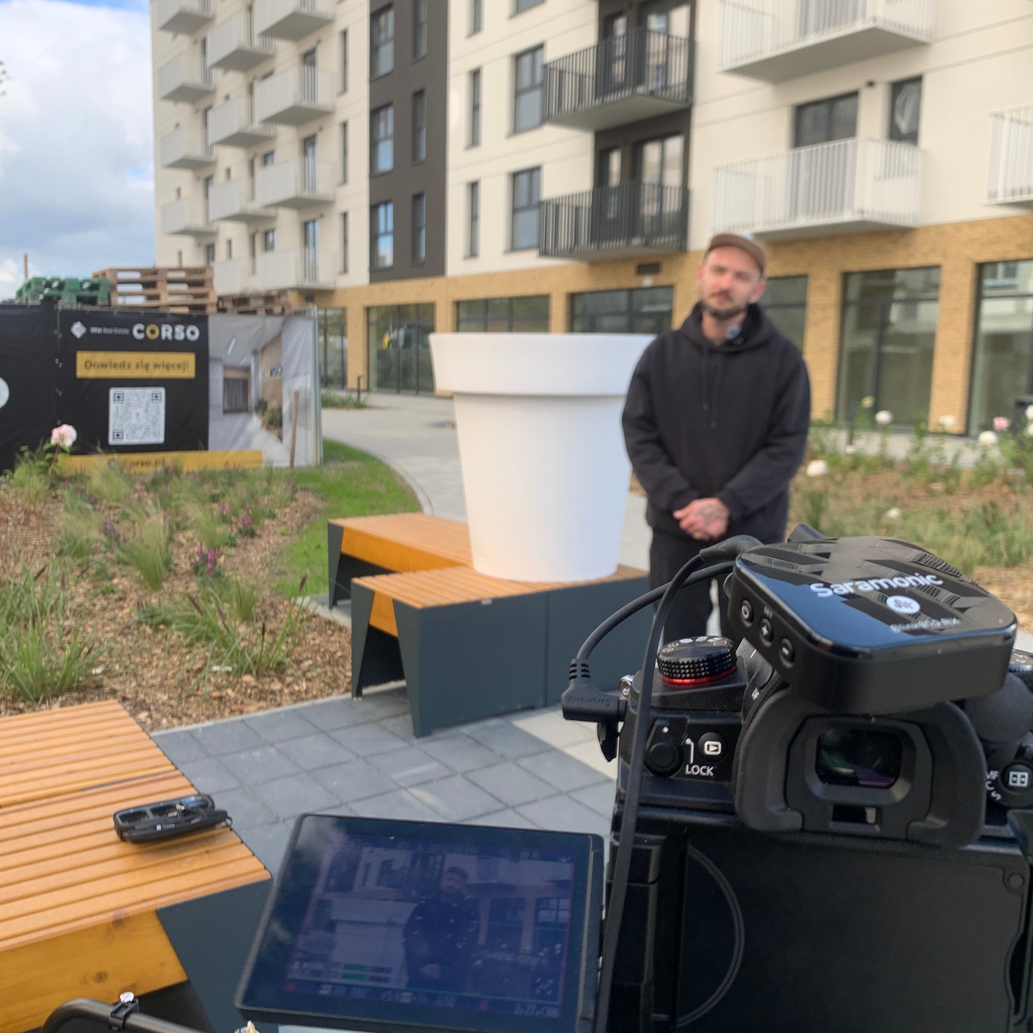 Kamera z mikrofonem Saramonic nagrywa mężczyznę na tle nowego osiedla z dużą donicą. Widoczny ekran kamery z podglądem nagrywanego materiału.