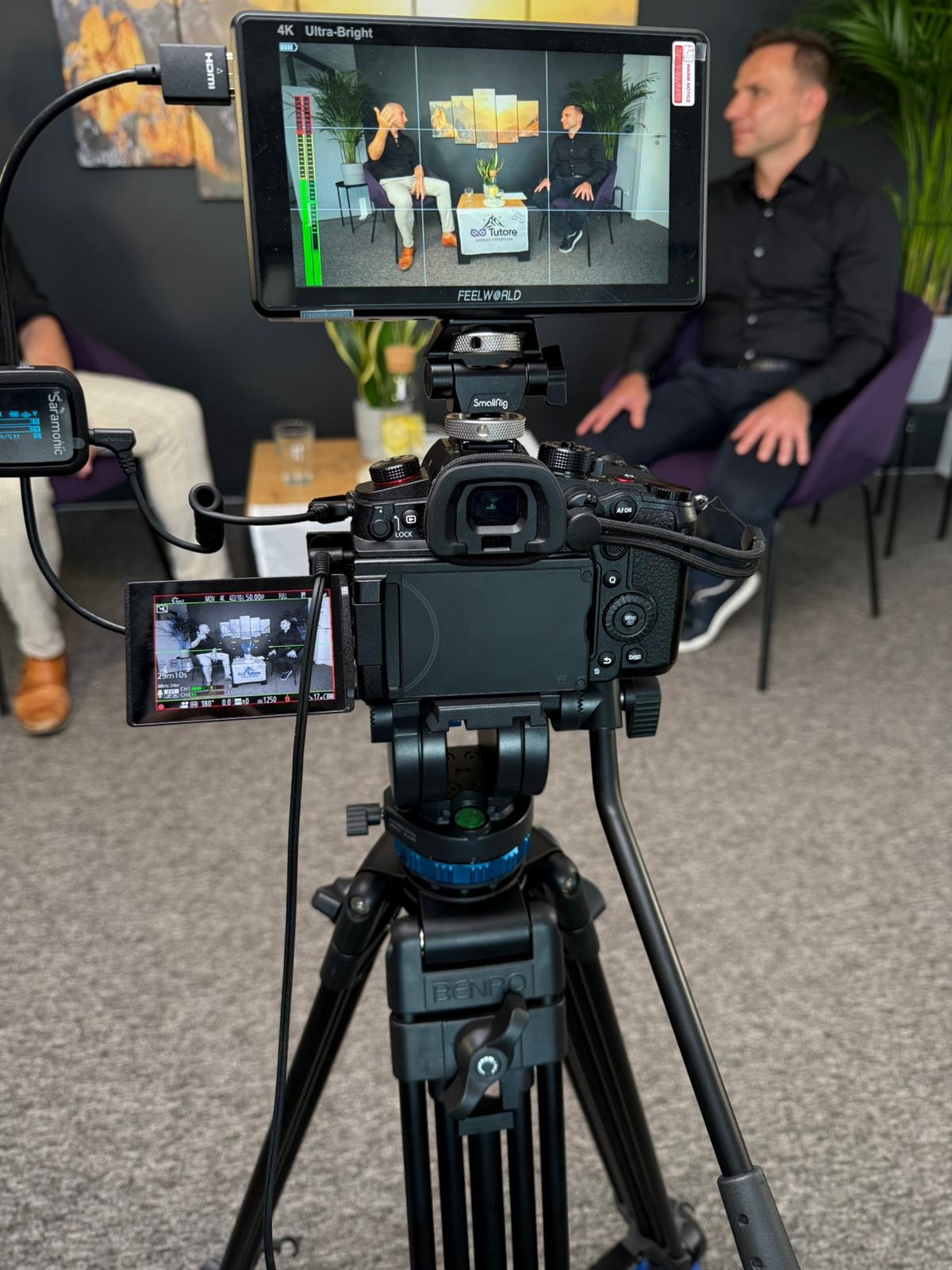 Kamera na statywie nagrywa wywiad z dwoma mężczyznami widocznymi na monitorze. Widoczny mikrofon Saramonic i monitor Feelworld 4K Ultra-Bright.