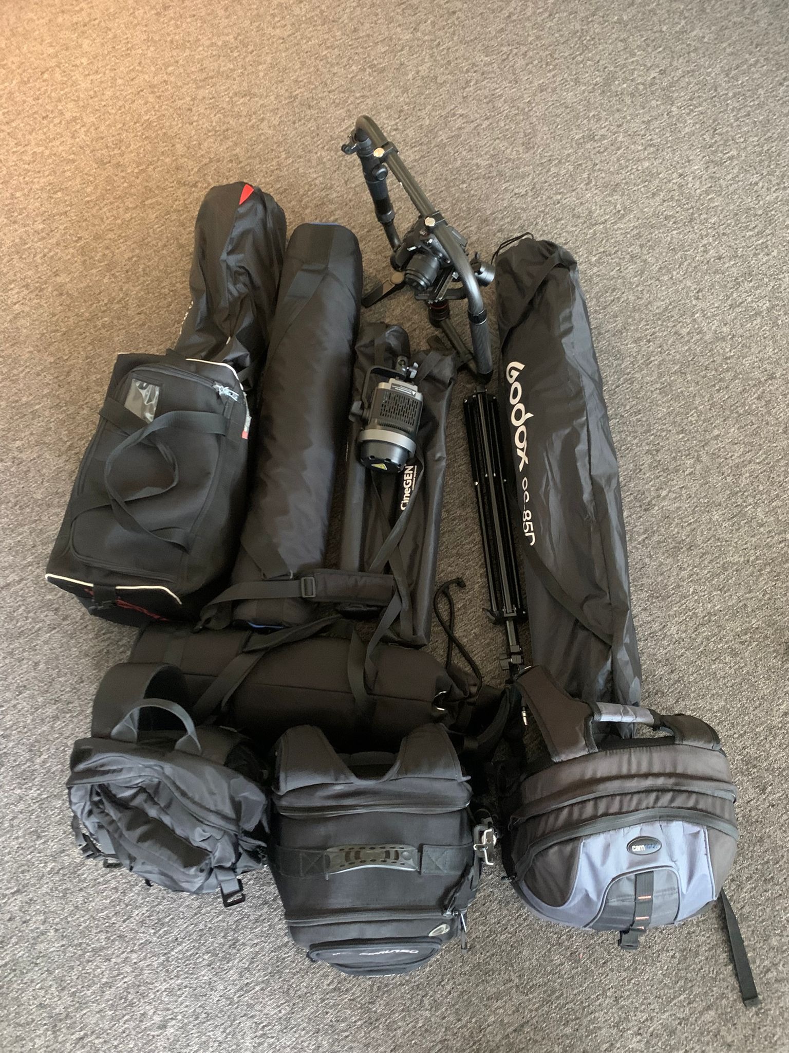 Sprzęt filmowy i fotograficzny: torby, statywy, oświetlenie CineGEN i Godox, rozłożone na szarym dywanie. Widok z góry, neutralne oświetlenie.