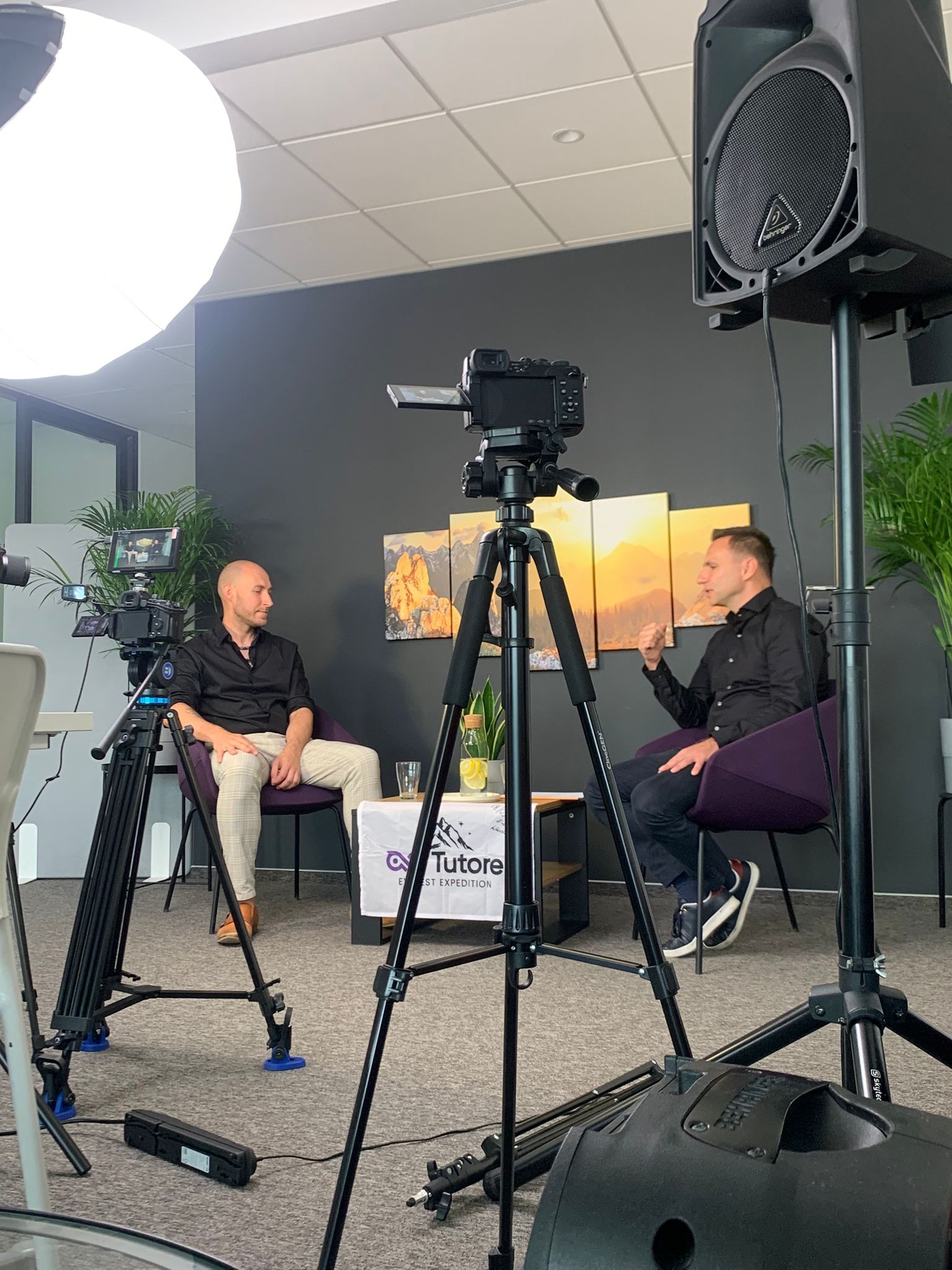 Dwie osoby w studio podczas nagrywania filmu, widoczne kamery na statywach, oświetlenie i głośnik. Na stole woda z cytryną i logo firmy.
