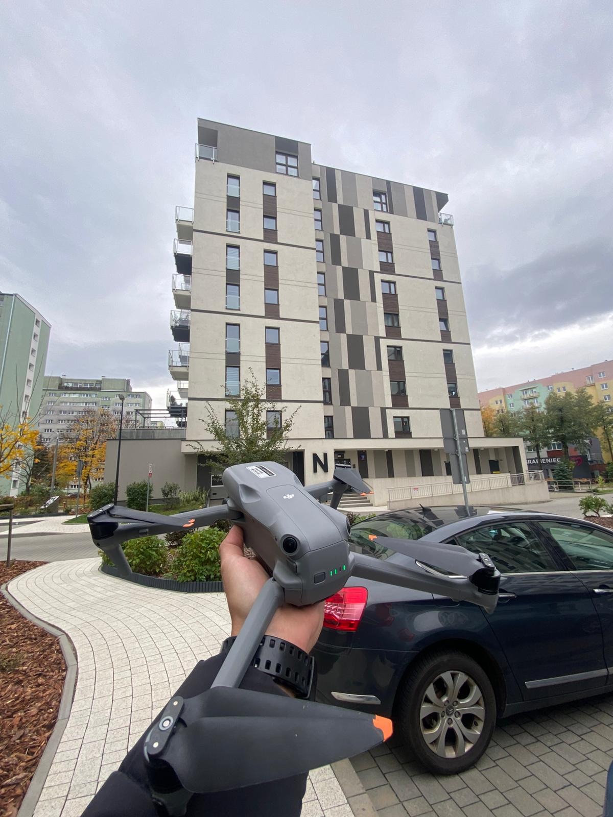 Szary dron DJI Mavic 3 w dłoni, na tle nowoczesnego budynku mieszkalnego i zaparkowanego samochodu. Ujęcie z dołu, pochmurne niebo.