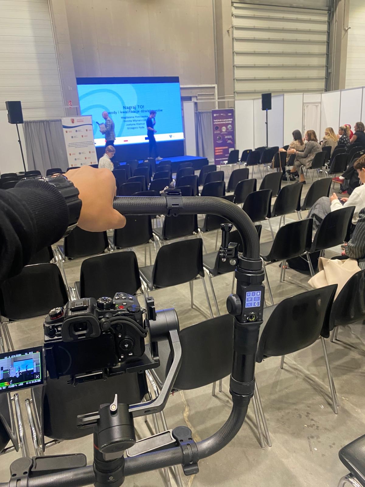 Kamera na stabilizatorze nagrywa prelekcję na tle rzędu krzeseł i sceny z ekranem. Ujęcie z perspektywy operatora, widoczny fragment ręki. Sala konferencyjna.