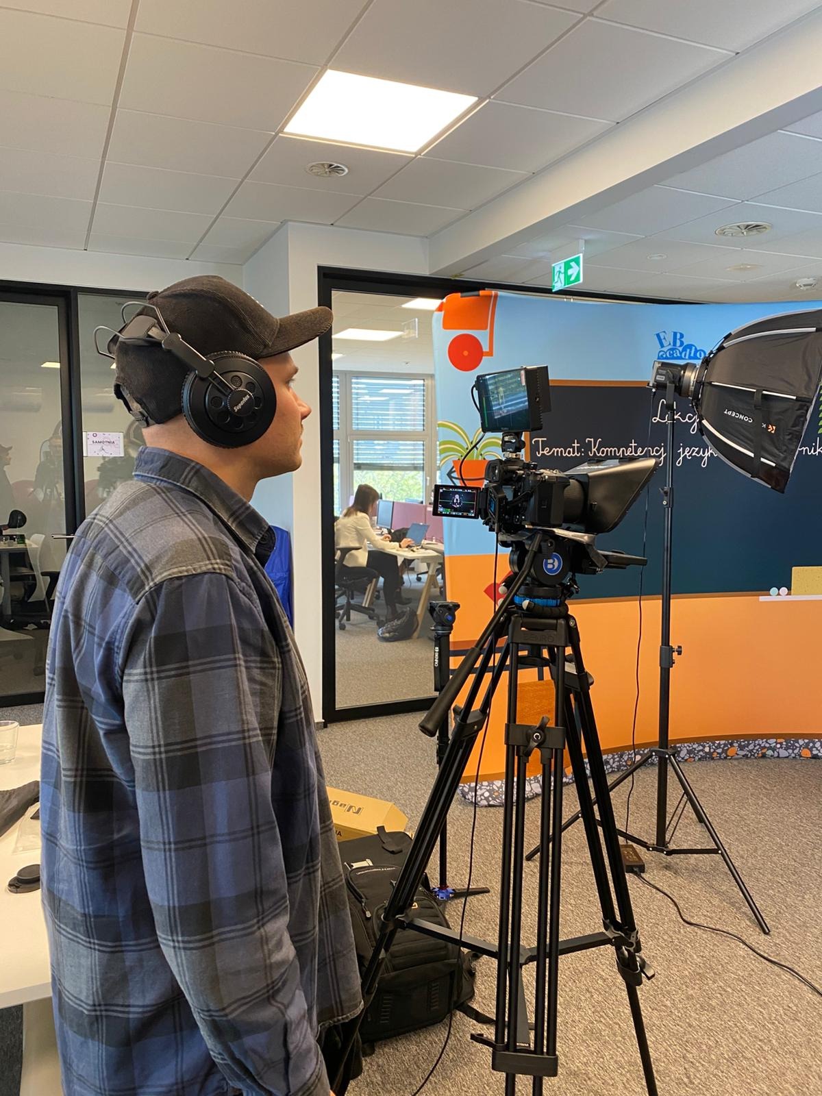 Kamerzysta w trakcie nagrywania lekcji z teleprompterem i oświetleniem studyjnym. Widoczny operator w słuchawkach i kamera na statywie. W tle biuro.