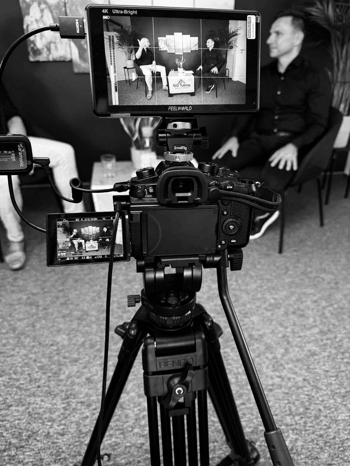 Kamera na statywie rejestruje wywiad z dwoma osobami. Widoczny monitor podglądowy z logo 'Q&S Tutore'. Czarno-biała fotografia z bliska, ukazująca sprzęt filmowy.
