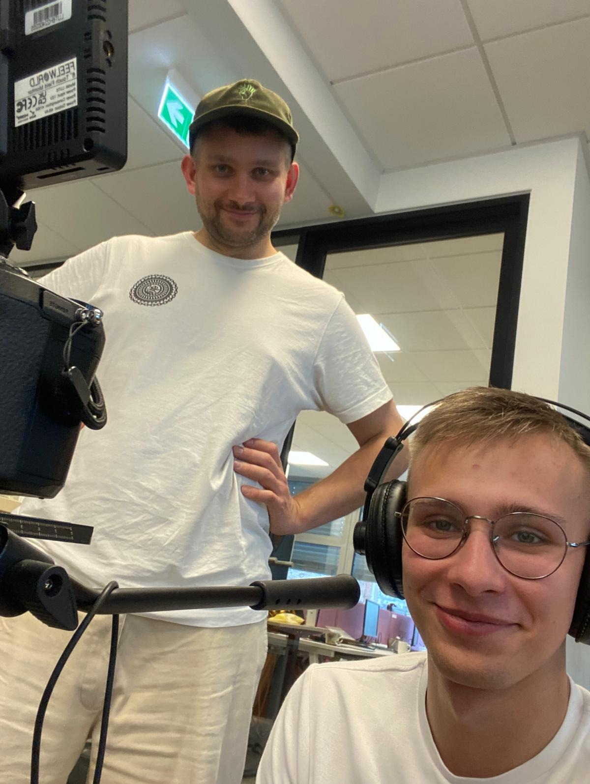 Ekipa filmowa w trakcie pracy: operator kamery z słuchawkami i drugi mężczyzna w tle, obaj w białych koszulkach, w jasnym pomieszczeniu z kamerą.