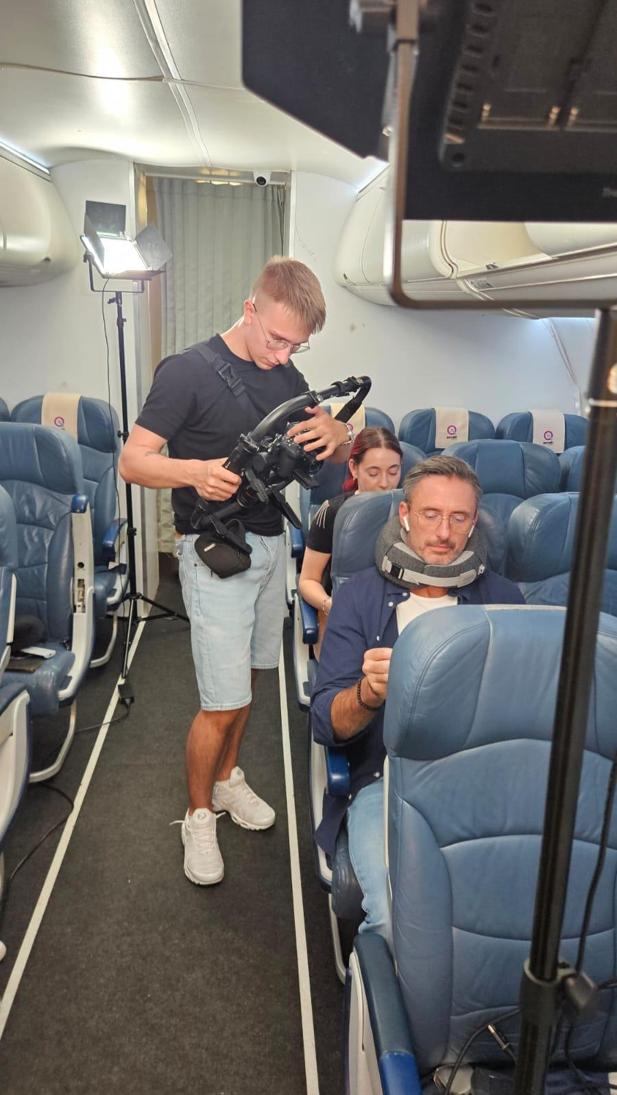 Kamerzysta z profesjonalnym sprzętem filmuje scenę wewnątrz samolotu. Aktor w podróżnej poduszce siedzi na fotelu. Dodatkowe oświetlenie.