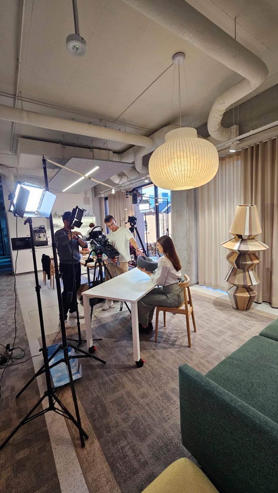 Ekipa filmowa kręci ujęcie z kobietą siedzącą przy stole w nowoczesnym wnętrzu. Widoczne kamery, oświetlenie i rekwizyty. Jasne, naturalne światło.