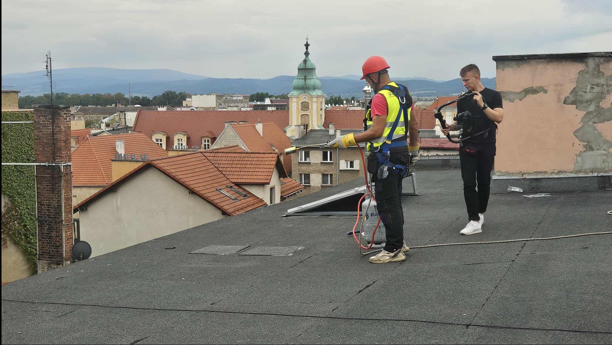 Ekipa na dachu: dekarz z palnikiem i kamerzysta filmujący proces. W tle panorama miasta z kościołem. Praca na wysokościach, zabezpieczenia, profesjonalny sprzęt.