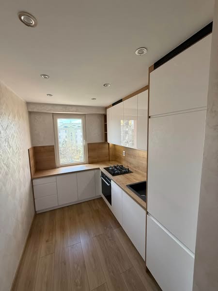 Nowoczesna kuchnia w kształcie litery L z białymi frontami i drewnianym blatem, oświetlona światłem dziennym wpadającym przez okno. Minimalistyczny design i funkcjonalne rozwiązania.