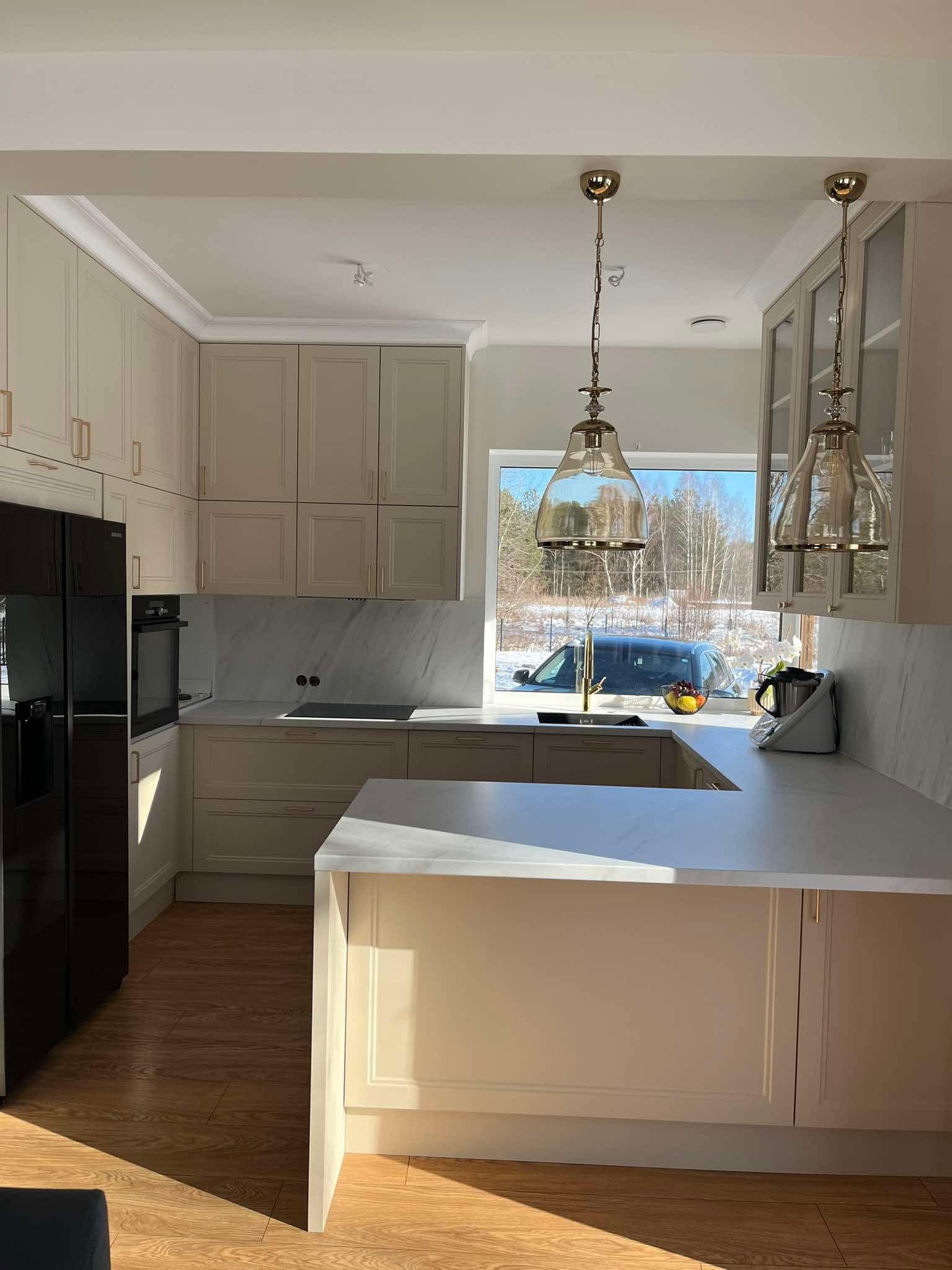 Jasna kuchnia w stylu klasycznym, z marmurowym blatem i złotymi detalami. Widoczne wiszące lampy i okno z widokiem na zimowy krajobraz. Eleganckie wykończenie.