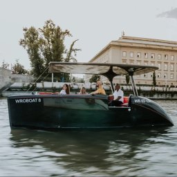 Skillmedia.pl - Rejs łodzią Wroboats po Odrze we Wrocławiu. Grupa osób na pokładzie, w tle Most Pokoju i budynek Uniwersytetu Wrocławskiego. Letnia sceneria.