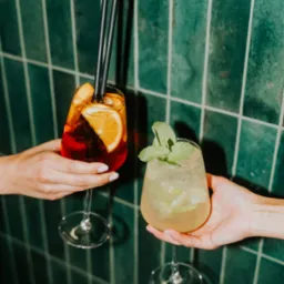 Dwa koktajle w kieliszkach trzymane w dłoniach na tle ściany z zielonych płytek. Jeden z pomarańczą i czarnymi słomkami, drugi z miętą.