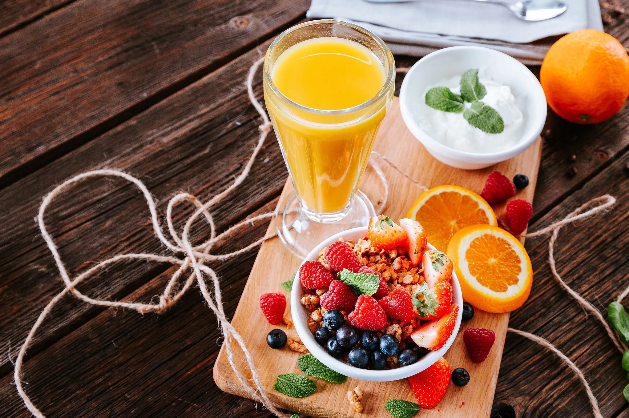 Kompozycja śniadaniowa na drewnianym stole: sok pomarańczowy, jogurt z miętą i miseczka z granolą, malinami, jagodami i truskawkami, podkreślona sznurkiem.