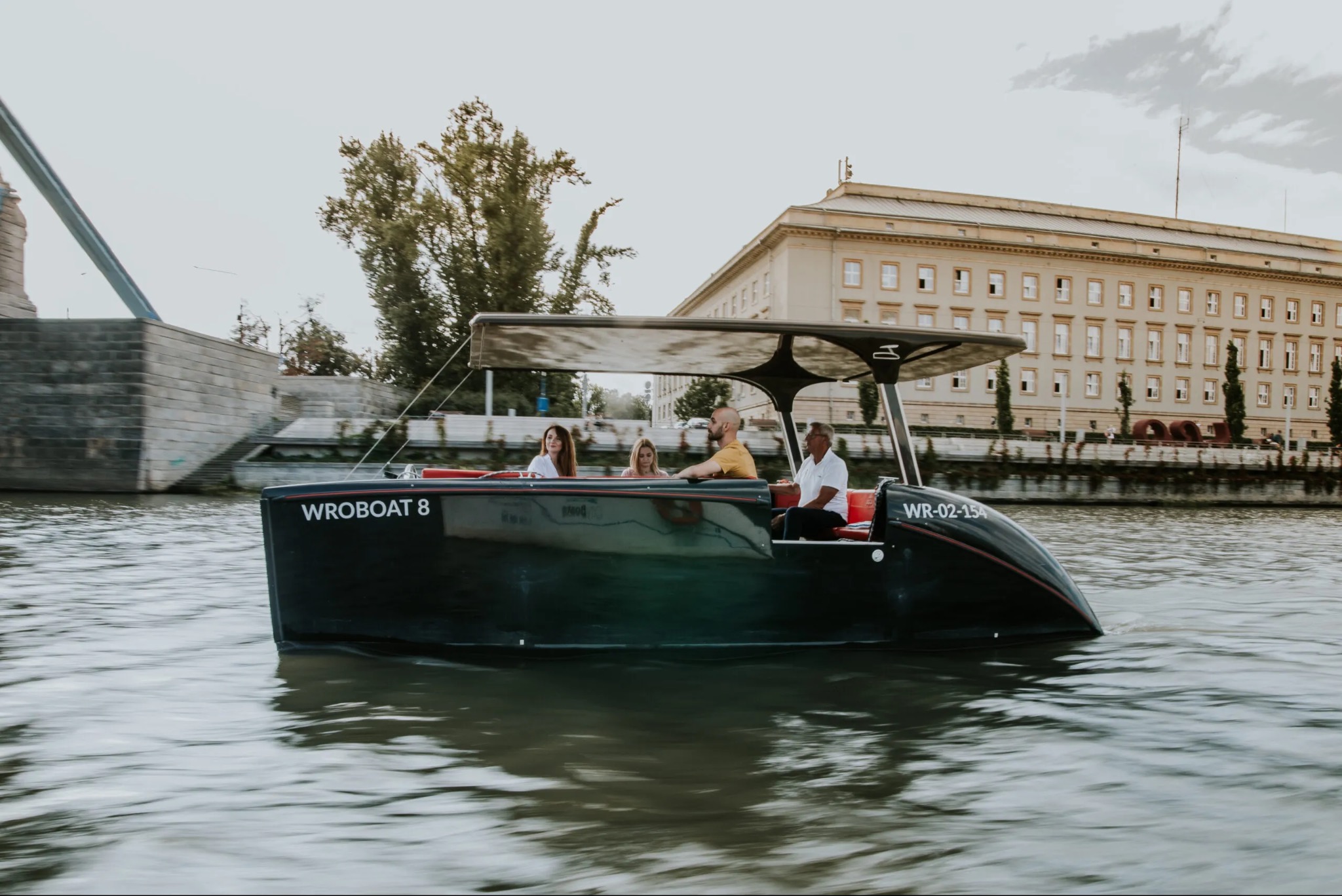 Rejs łodzią Wroboats po Odrze we Wrocławiu. Grupa osób na pokładzie, w tle Most Pokoju i budynek Uniwersytetu Wrocławskiego. Letnia sceneria.