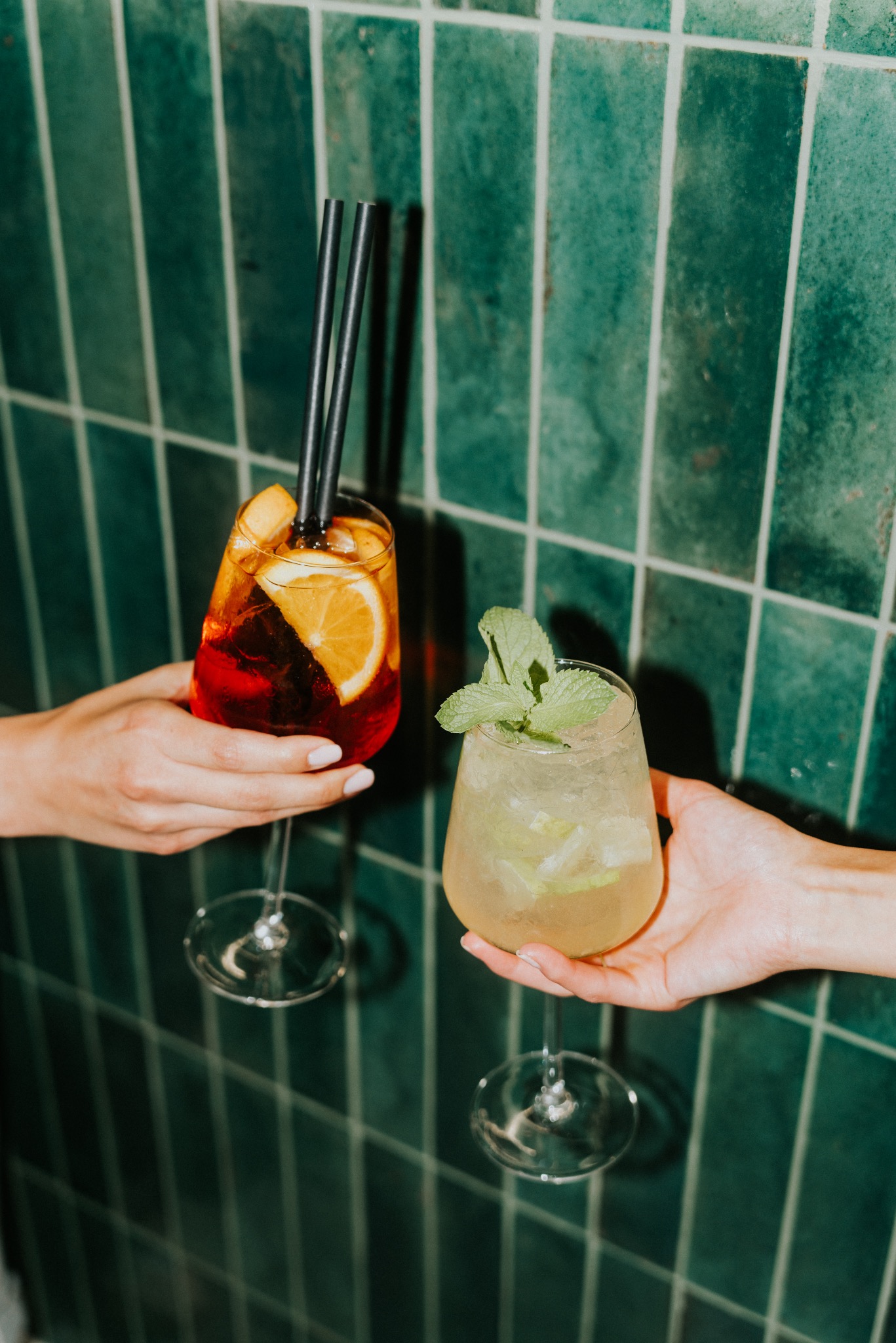 Dwa koktajle w kieliszkach trzymane w dłoniach na tle ściany z zielonych płytek. Jeden z pomarańczą i czarnymi słomkami, drugi z miętą.