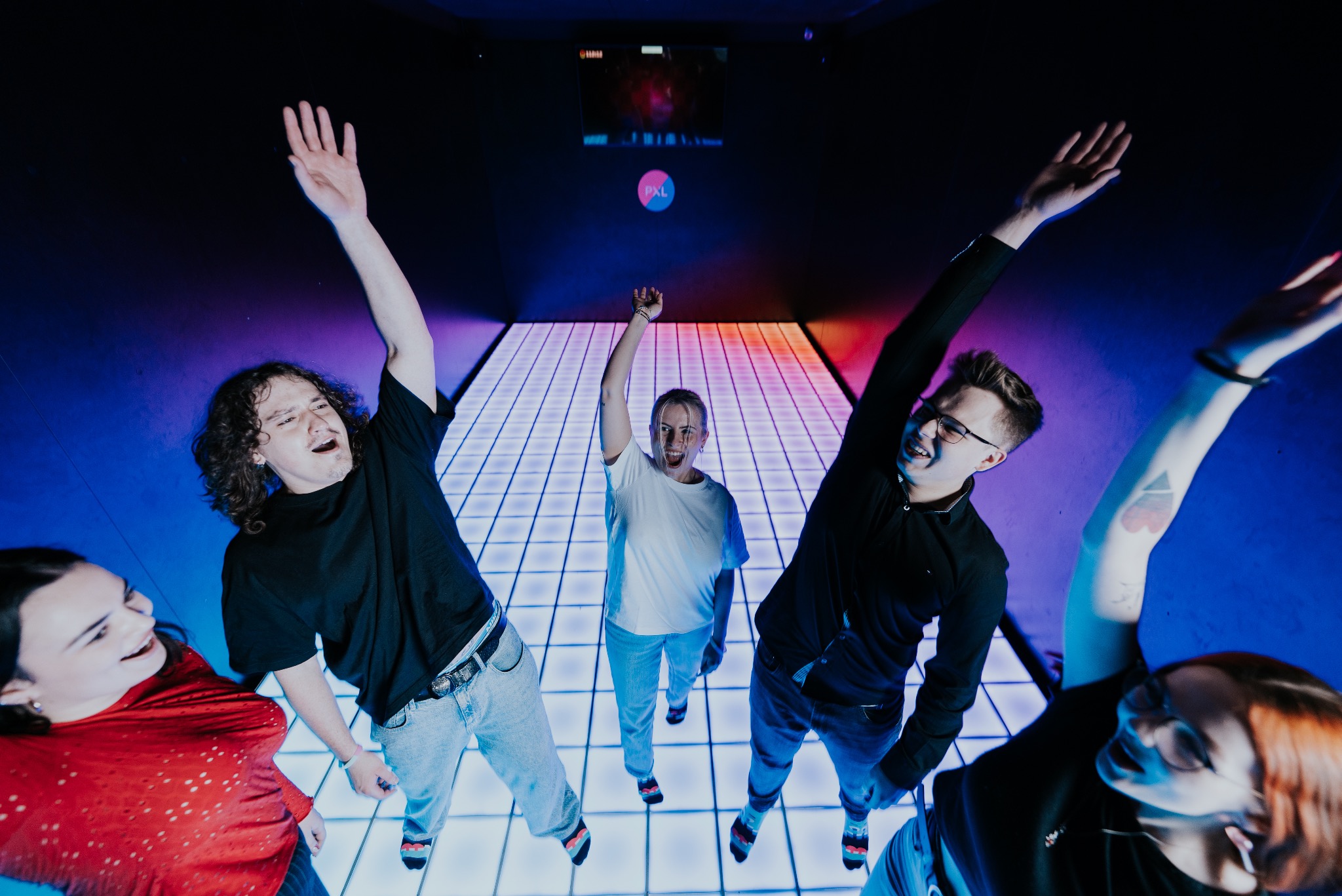 Grupa radosnych osób z podniesionymi rękami tańczy na podświetlanym parkiecie w klubie. Kolorowe światła tworzą dynamiczną atmosferę. Widoczny ekran w tle.