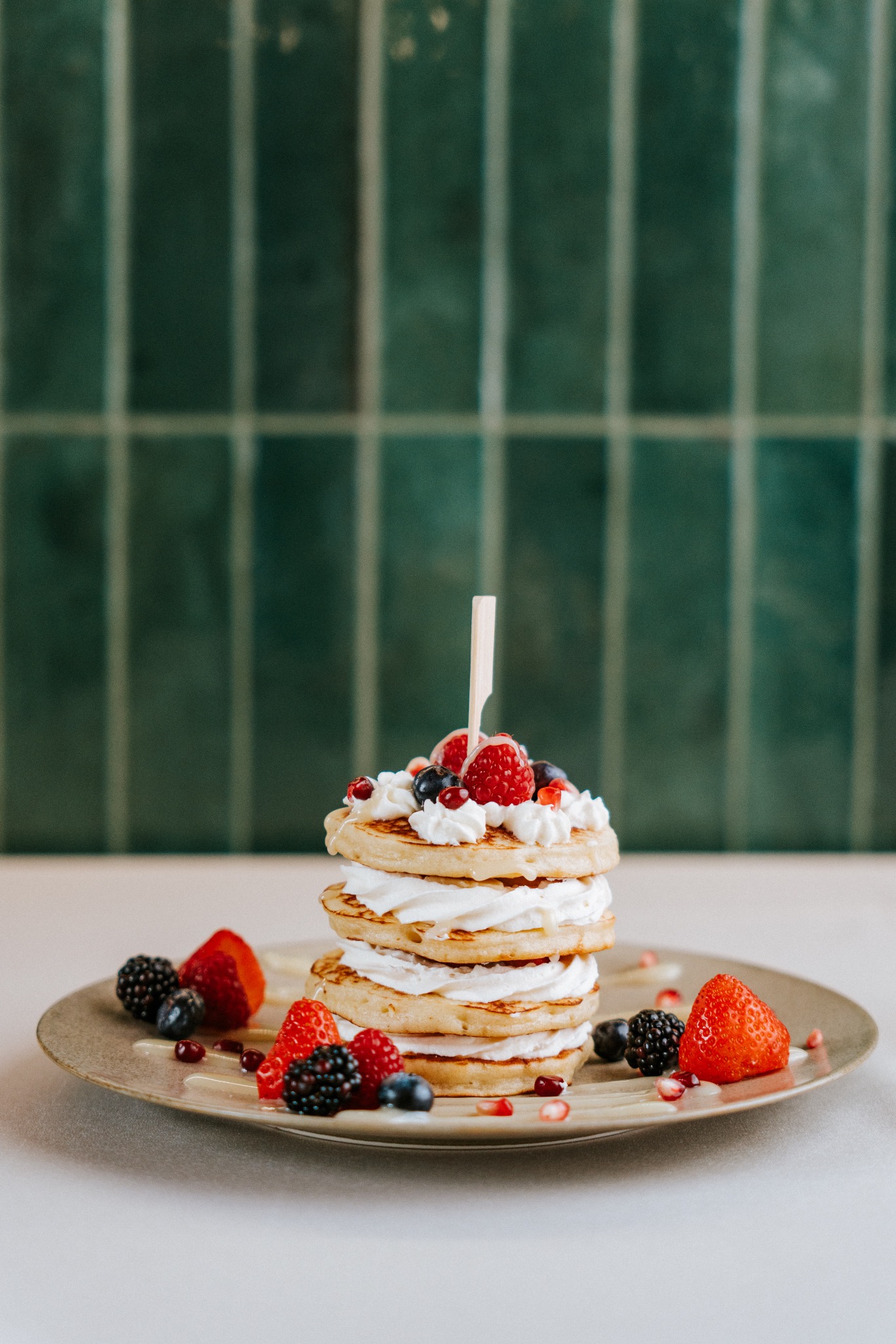 Apetyczne pancakes z bitą śmietaną i świeżymi owocami (maliny, jagody, jeżyny) na talerzu, na tle zielonej ściany z płytek. Słodki deser w zbliżeniu.