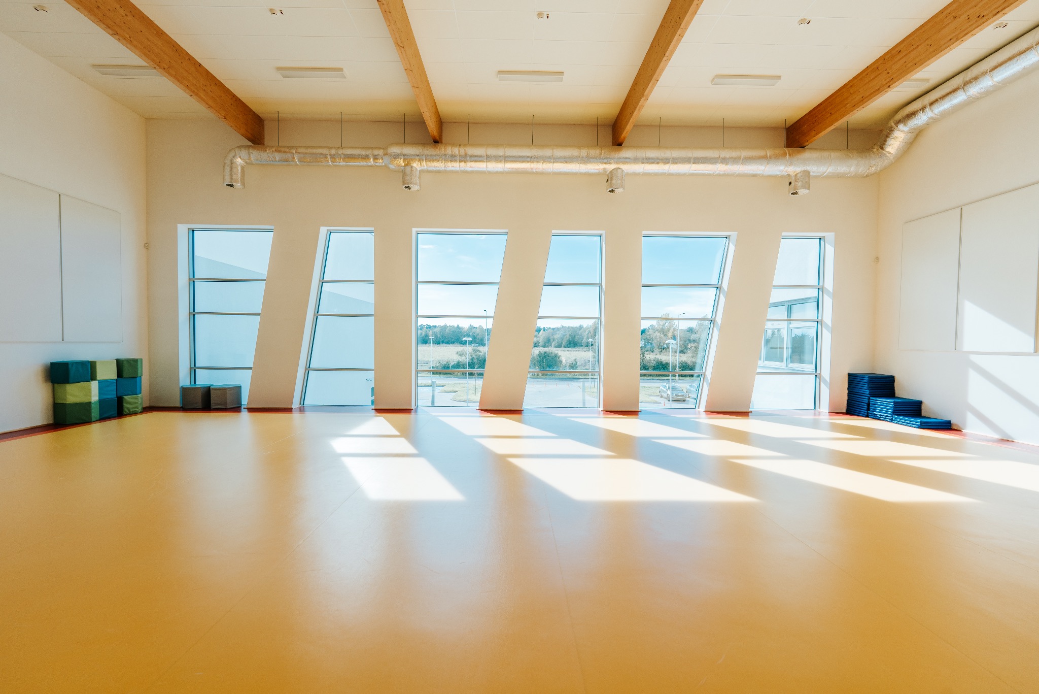 Jasne wnętrze sali gimnastycznej z żółtą podłogą, nietypowymi oknami i drewnianymi belkami na suficie. Widoczne maty do ćwiczeń i wentylacja.