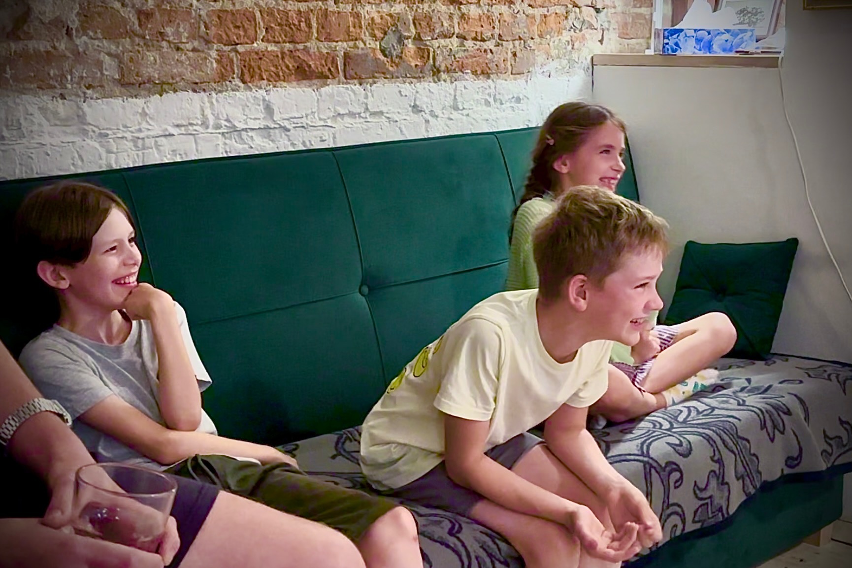 Uśmiechnięte dzieci siedzą na zielonej kanapie przed ceglaną ścianą, prawdopodobnie podczas imprezy lub oglądania występu.