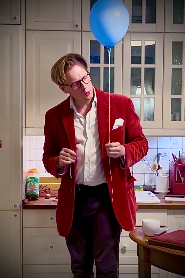 Mężczyzna w czerwonej marynarce i okularach trzyma niebieski balon w kuchni, sugerując występ lub event.