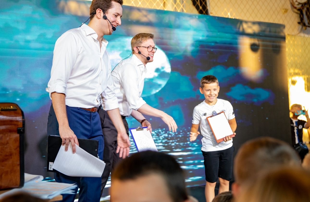 Dwóch konferansjerów z mikrofonami prezentuje występ iluzjonistyczny z udziałem chłopca na tle ekranu z motywem księżyca i chmur.