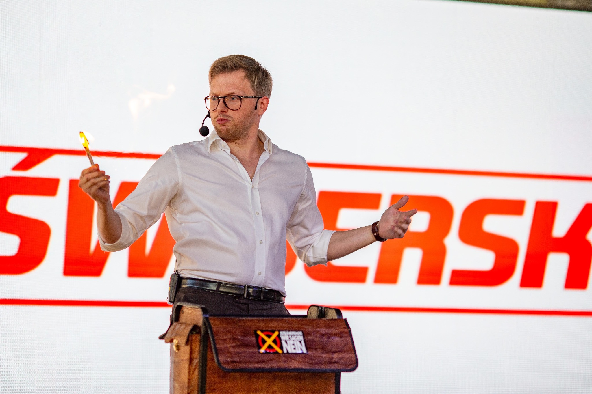 Iluzjonista w trakcie występu. Mężczyzna w okularach i białej koszuli trzyma płonącą pałeczkę na tle dużego, czerwonego napisu. Na pierwszym planie brązowa skrzynia.