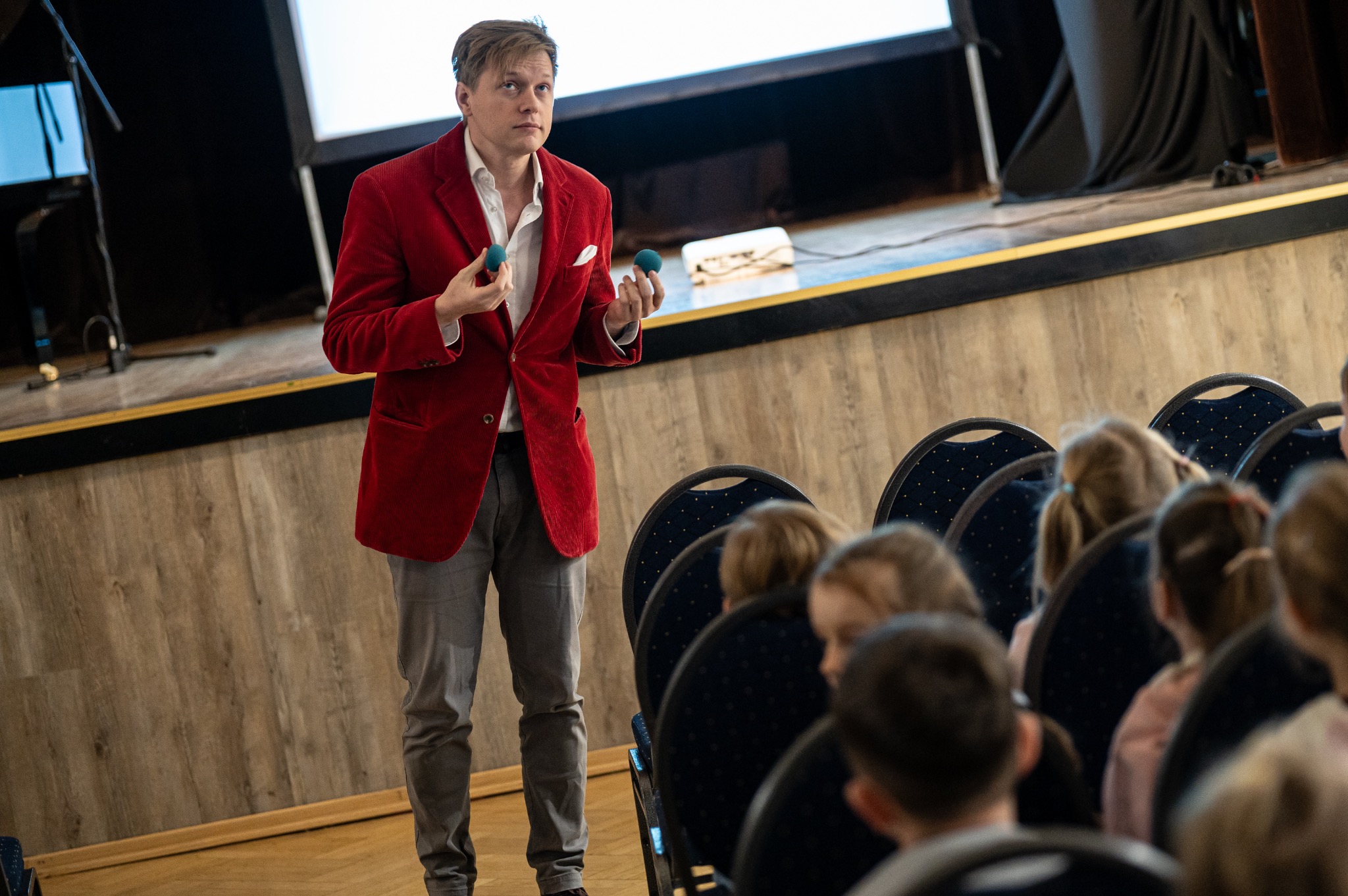 Młody mężczyzna w czerwonej marynarce prezentuje trik z zielonymi kulkami na scenie przed publicznością. Scena z drewnianym wykończeniem, rzędy krzeseł.