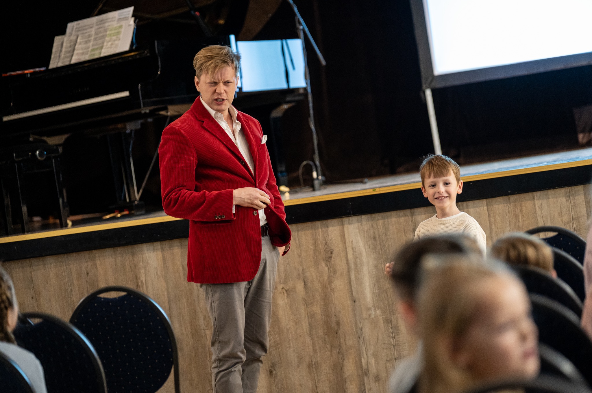 Iluzjonista w czerwonej marynarce na scenie z pianinem w tle, uśmiechnięty chłopiec w tłumie. Pokaz iluzji dla dzieci.