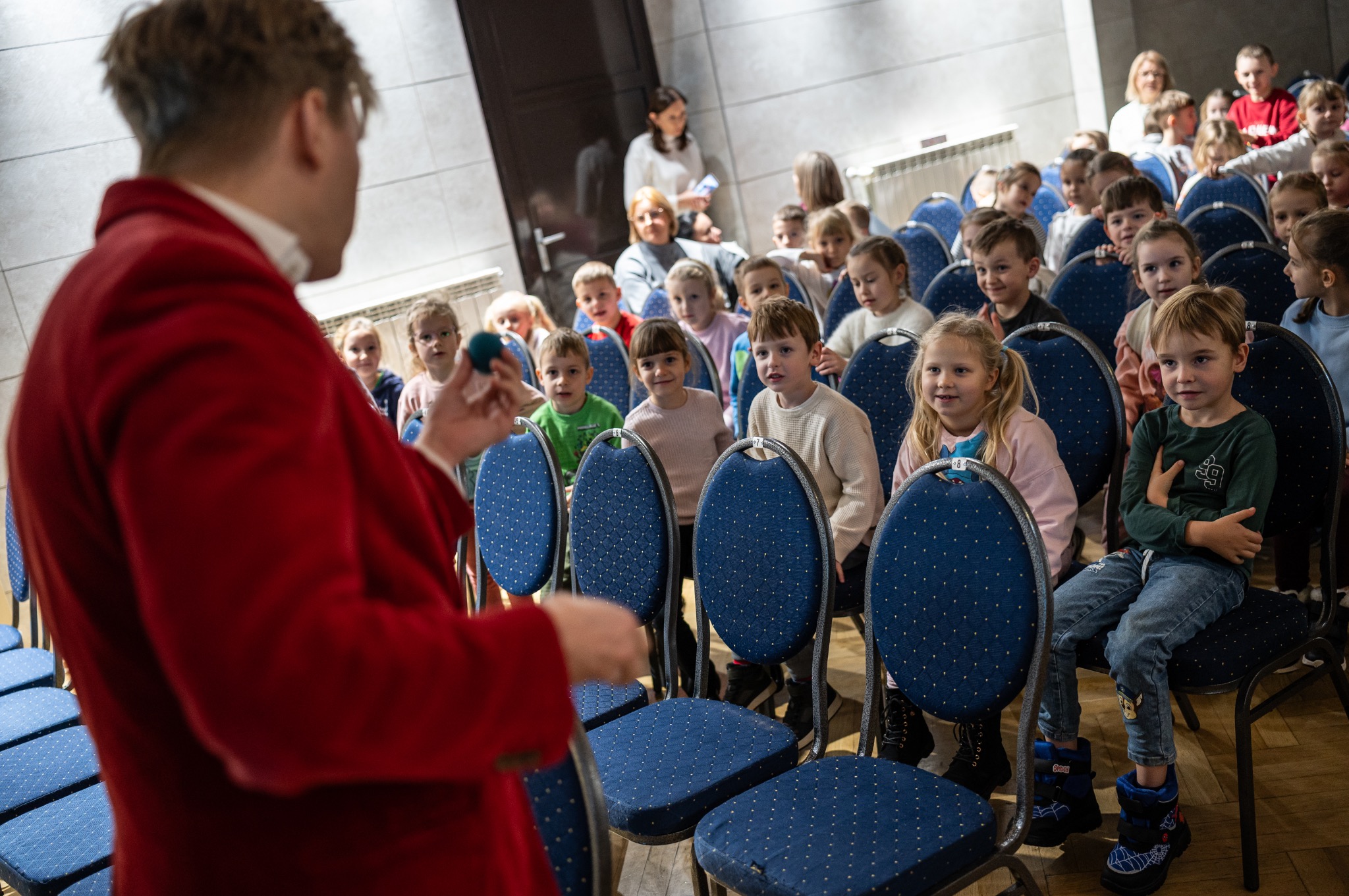 Występ artysty w czerwonym fraku przed rzędem zaciekawionych dzieci siedzących na niebieskich krzesłach podczas pokazu iluzji z małą, zieloną kulką.