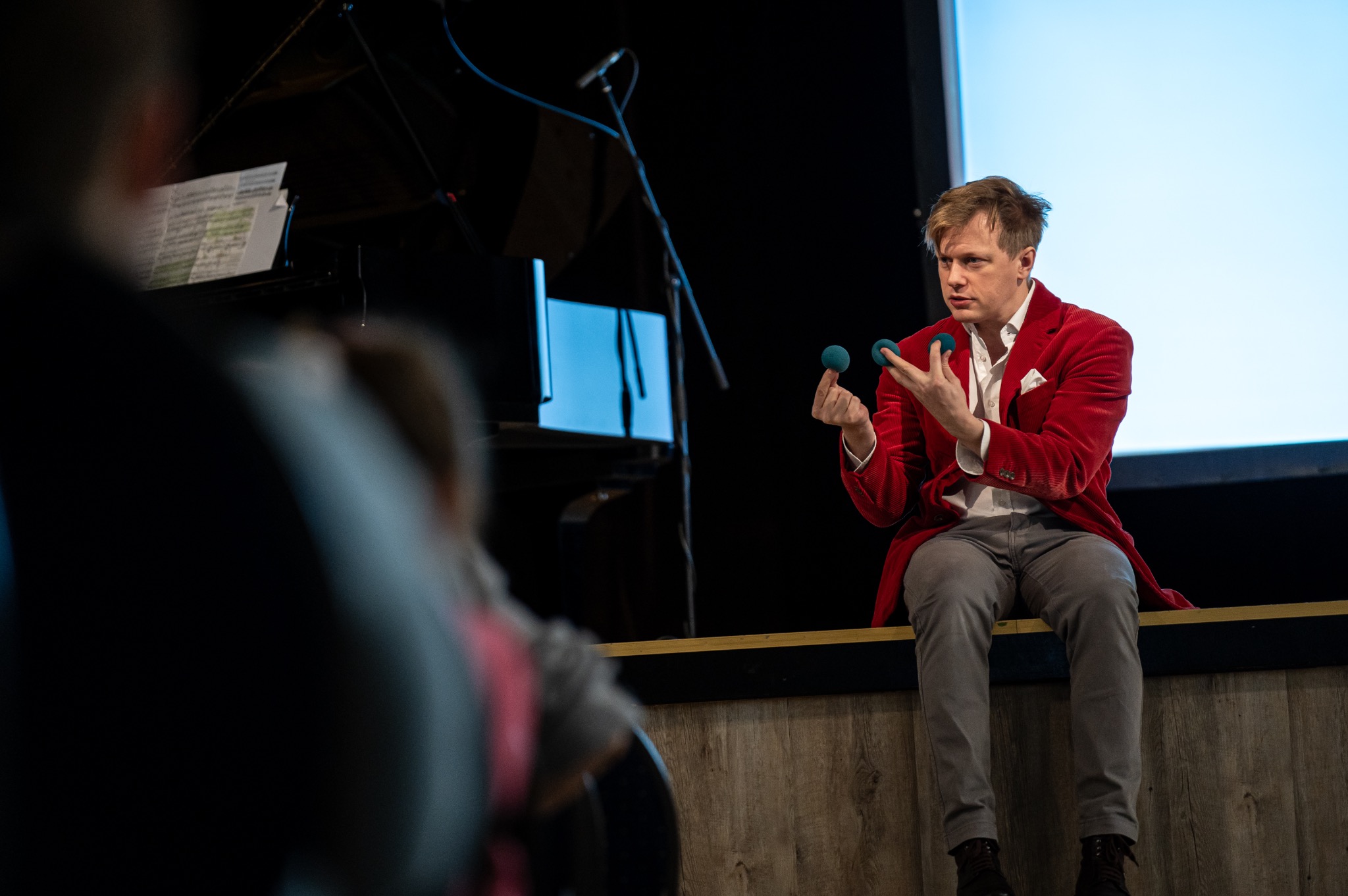 Mężczyzna w czerwonej marynarce prezentuje trik z zielonymi kulkami na scenie. W tle pianino i publiczność. Scena oświetlona, z widocznym ekranem w tle.