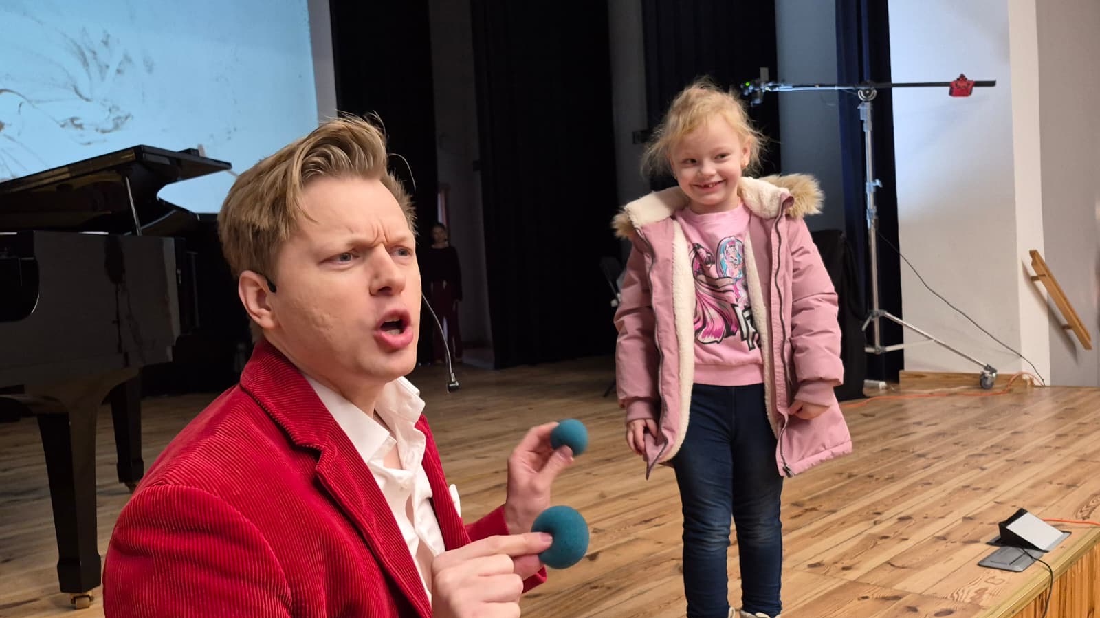 Występ iluzjonisty w czerwonej marynarce z rekwizytami na scenie, obok uśmiechnięta dziewczynka w różowej kurtce. W tle pianino i oświetlenie sceniczne.