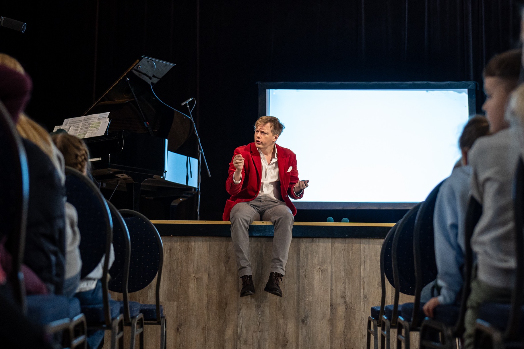 Artysta w czerwonej marynarce siedzi na scenie przed publicznością, gestykulując podczas występu. W tle fortepian i ekran projekcyjny.