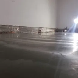 Świeżo wylana, mokra powierzchnia posadzki w hali, zabezpieczona folią przy ścianach. Widoczne odbicia światła i pracownicy w tle.