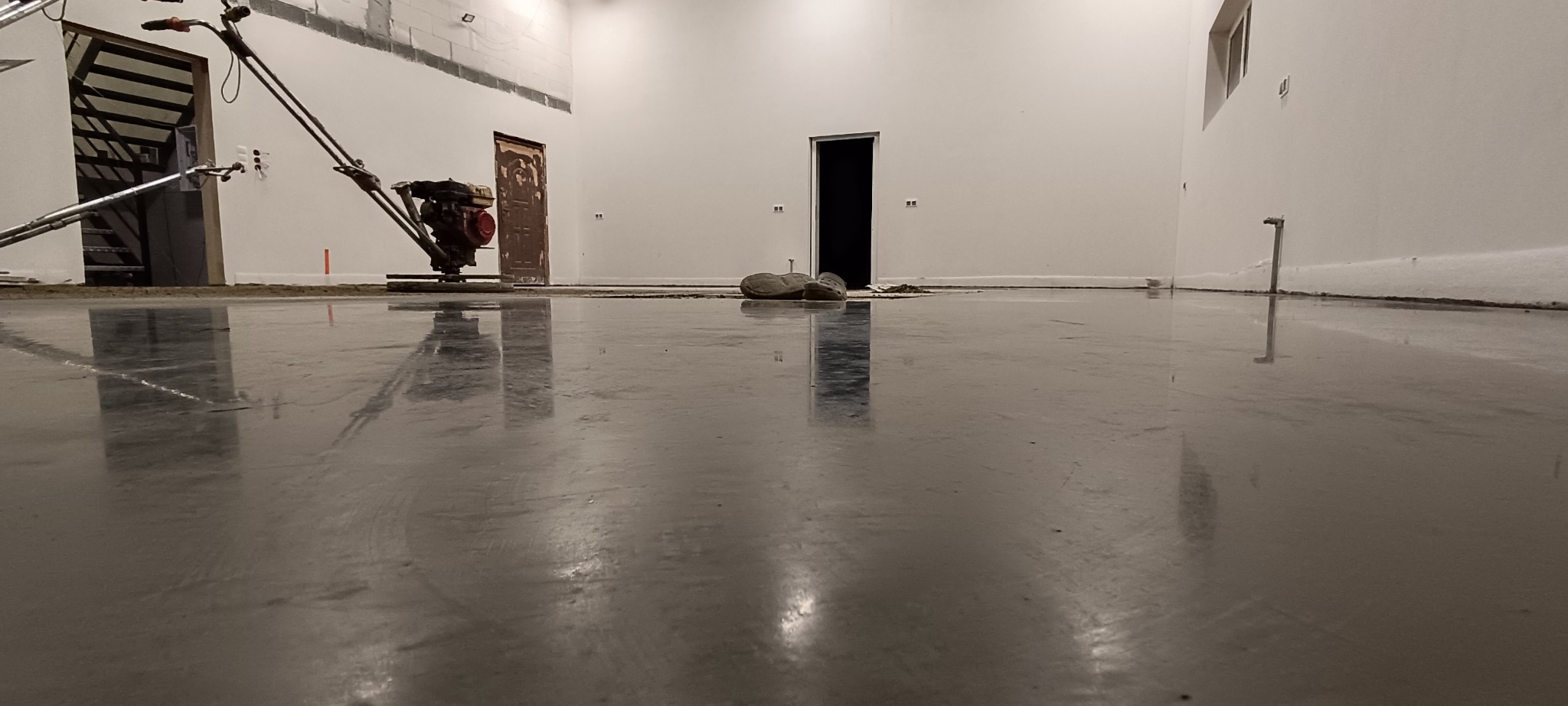 Świeżo wylana, gładka posadzka przemysłowa z maszyną do zacierania w tle. Widoczne odbicia w mokrej powierzchni i niedokończone ściany. Buty robocze leżą obok drzwi.