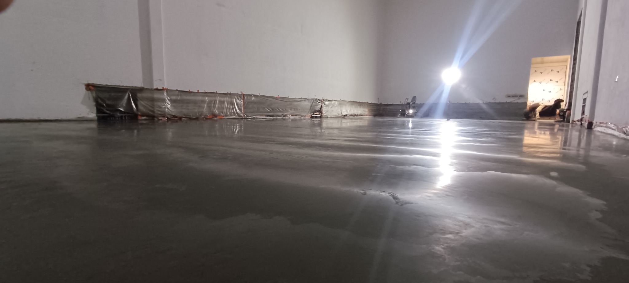 Świeżo wylana, mokra powierzchnia posadzki w hali, zabezpieczona folią przy ścianach. Widoczne odbicia światła i pracownicy w tle.