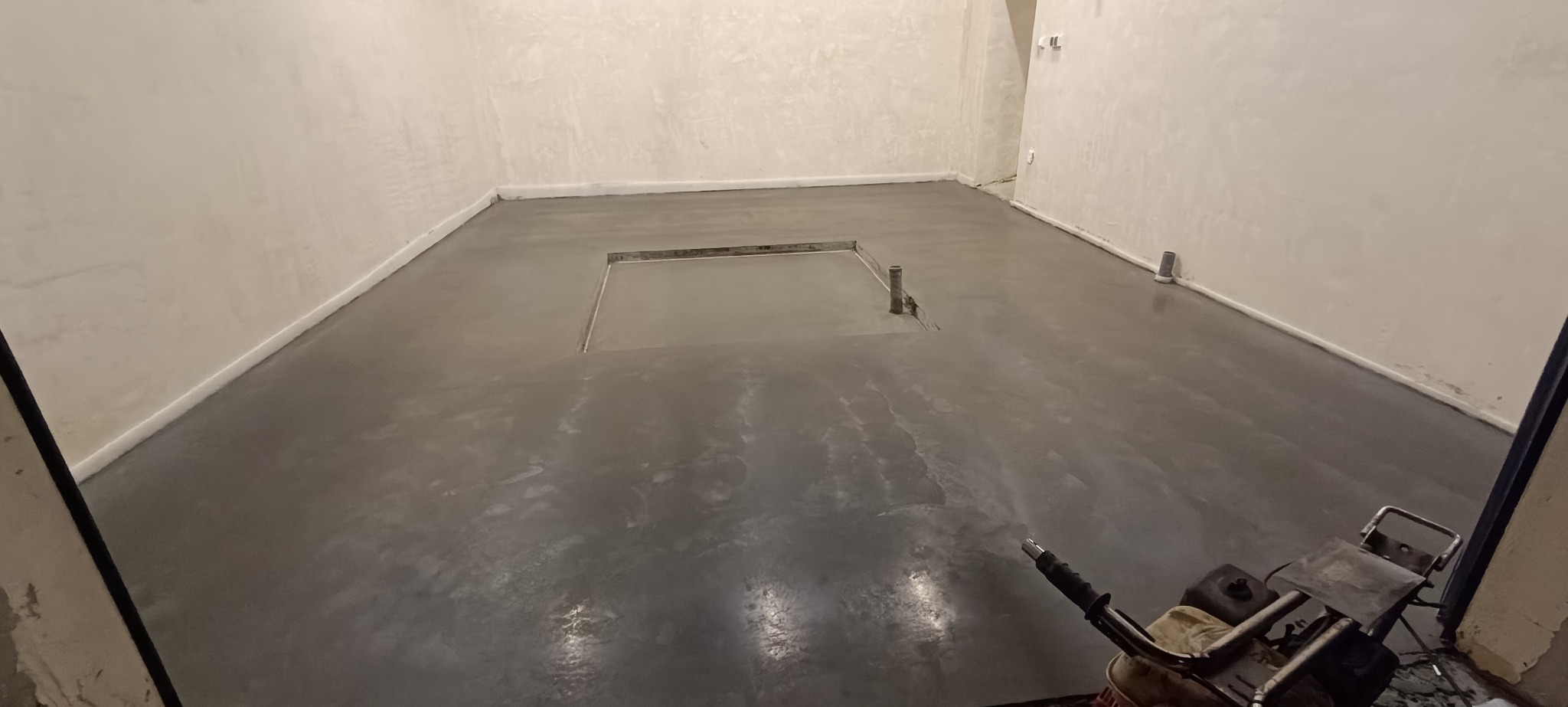 Świeża, gładka wylewka w pomieszczeniu z widocznym włazem i wystającymi rurami. Na pierwszym planie fragment maszyny do wygładzania betonu.