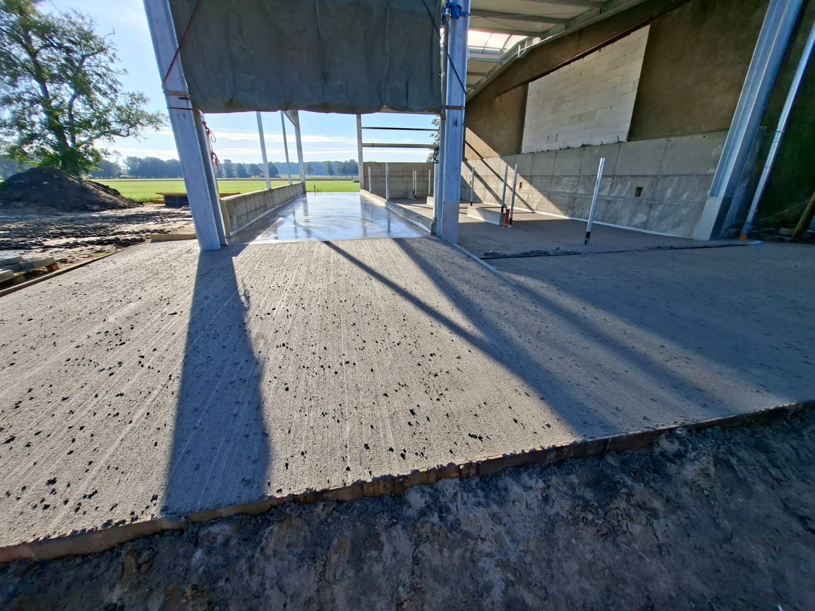 Świeżo wylana, szara powierzchnia betonowa wewnątrz konstrukcji hali, z widocznymi cieniami słupów i fragmentem zielonego krajobrazu w tle.