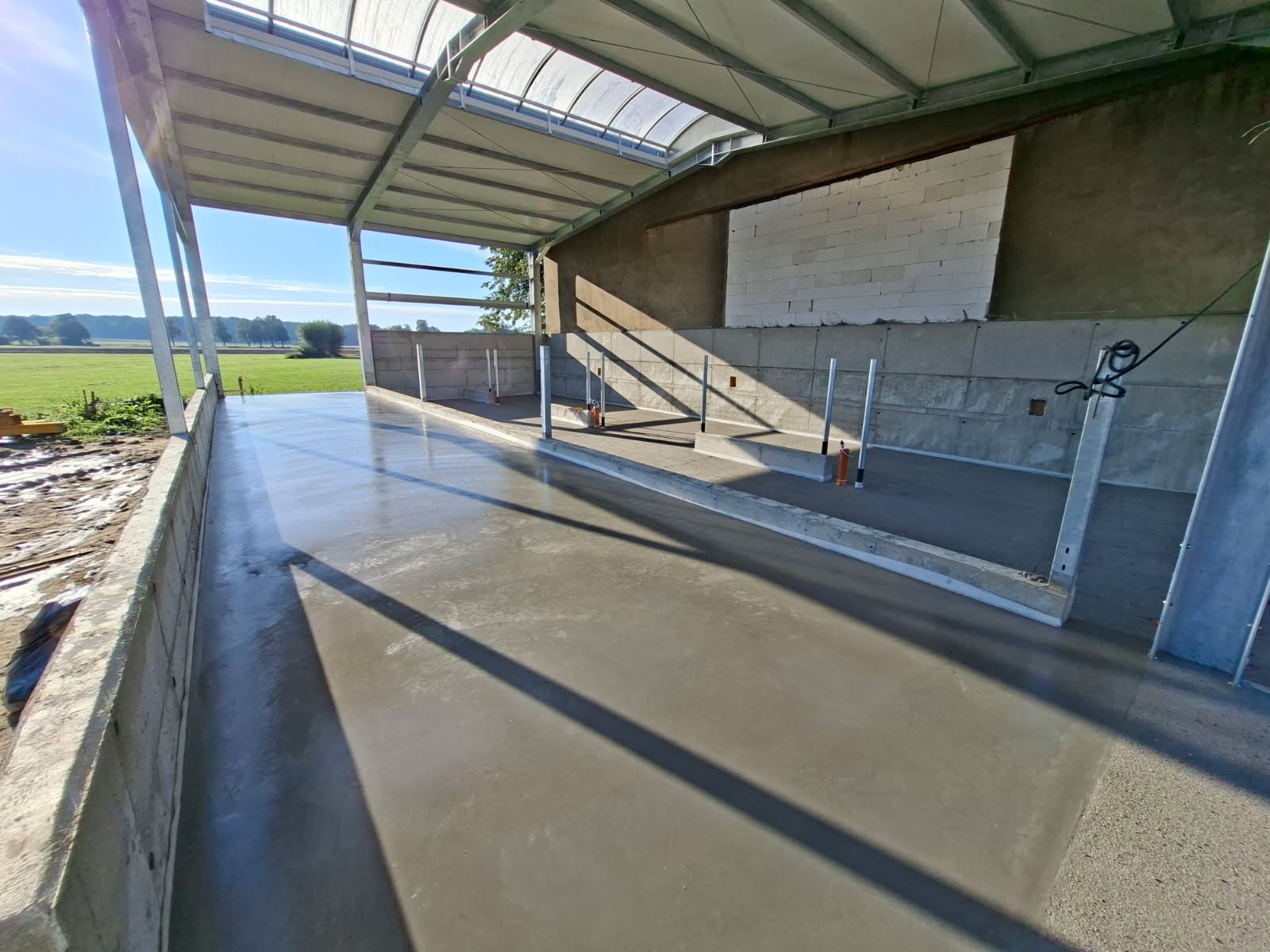 Świeżo wylana, gładka wylewka w hali z konstrukcją stalową i betonowymi ścianami. Widoczne pionowe słupki i cień padający na podłogę. Na zewnątrz zielony krajobraz.