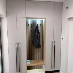 Krystian Zaręba - Biała szafa wnękowa z drewnianym panelem i oświetleniem LED, czarne uchwyty, drewniana podłoga. Wnęka z wieszakami i siedziskiem. Nowoczesny design.
