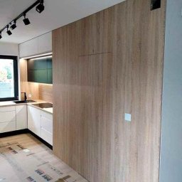 Krystian Zaręba - Nowoczesna kuchnia: białe szafki, drewniana ściana z ukrytymi drzwiami, czarne akcenty i oświetlenie punktowe. Podłoga zabezpieczona kartonem.