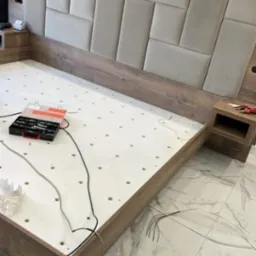 Montaż drewnianego ł&oacute;żka z tapicerowanym zagł&oacute;wkiem w odcieniach beżu. Widoczne narzędzia i elementy konstrukcyjne. Praca w toku.