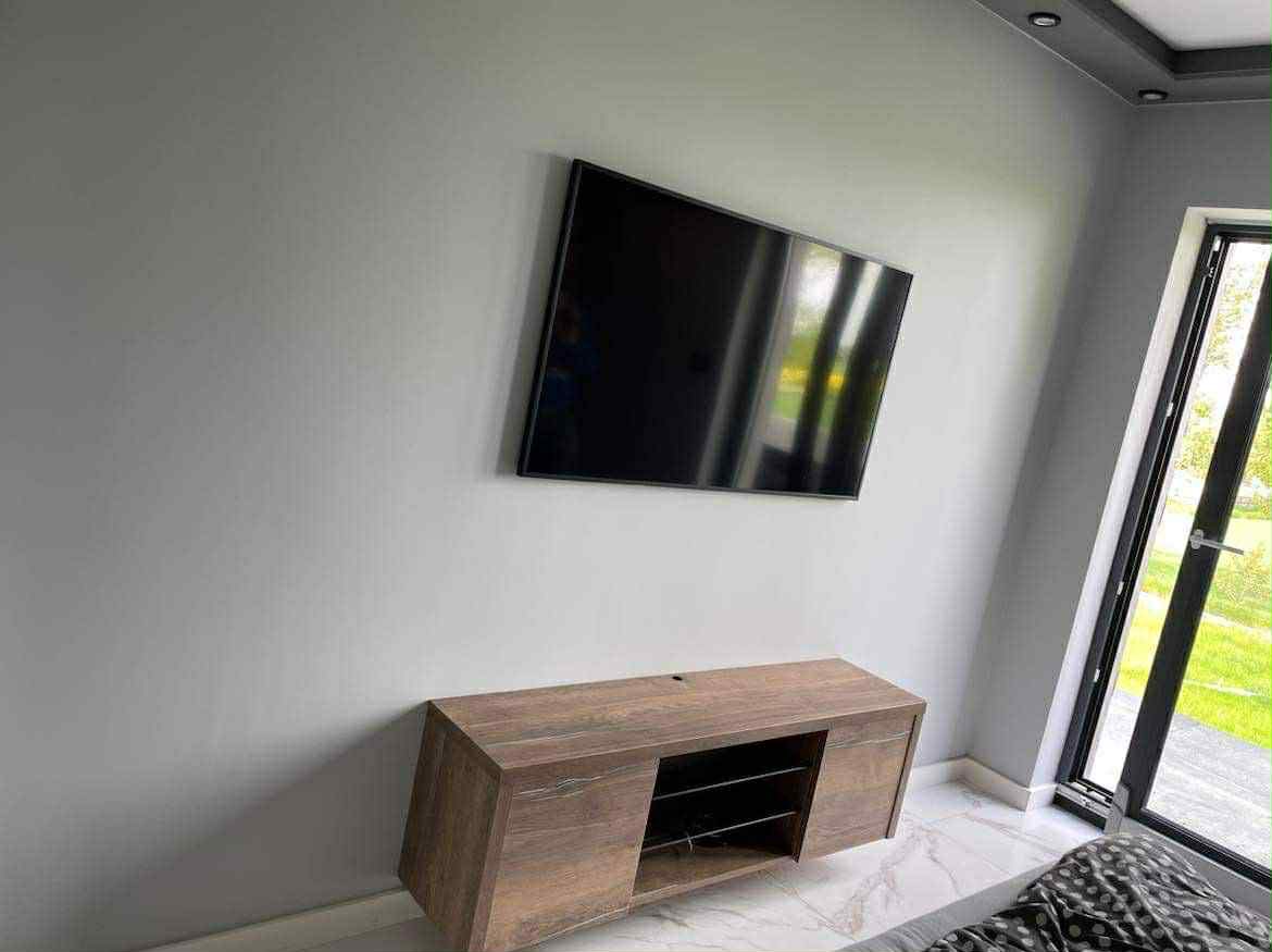 Nowoczesna szafka RTV z drewna na ścianie pod telewizorem, minimalistyczny design, jasne wnętrze z dużym oknem, marmurowe płytki podłogowe.