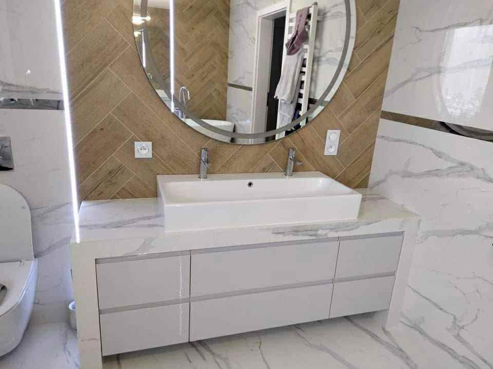 Nowoczesna łazienka z owalnym lustrem, białą umywalką nablatową na szafce z szufladami i marmurowymi płytkami. Uwagę przyciąga wzór jodełki na ścianie.