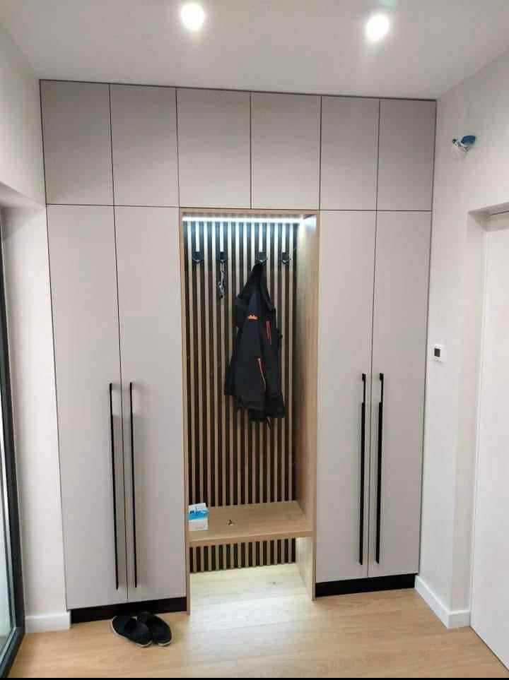 Biała szafa wnękowa z drewnianym panelem i oświetleniem LED, czarne uchwyty, drewniana podłoga. Wnęka z wieszakami i siedziskiem. Nowoczesny design.