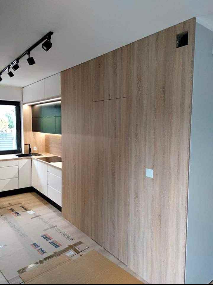 Nowoczesna kuchnia: białe szafki, drewniana ściana z ukrytymi drzwiami, czarne akcenty i oświetlenie punktowe. Podłoga zabezpieczona kartonem.