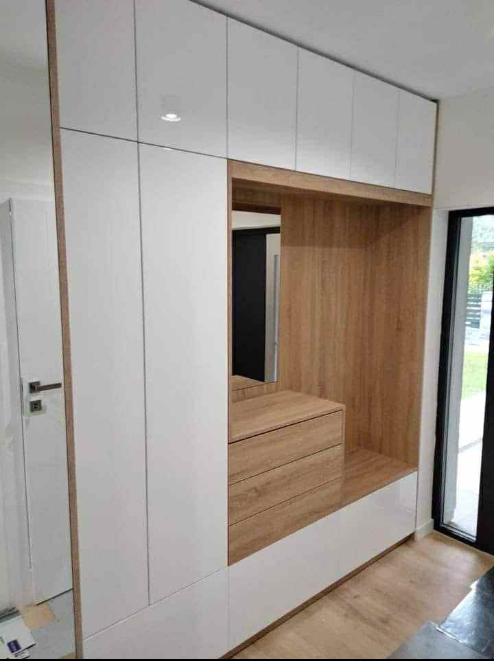 Nowoczesna szafa wnękowa z białymi frontami i drewnianymi akcentami, wbudowana w przedpokoju, z lustrem i szufladami. Minimalistyczny design, jasne wnętrze.