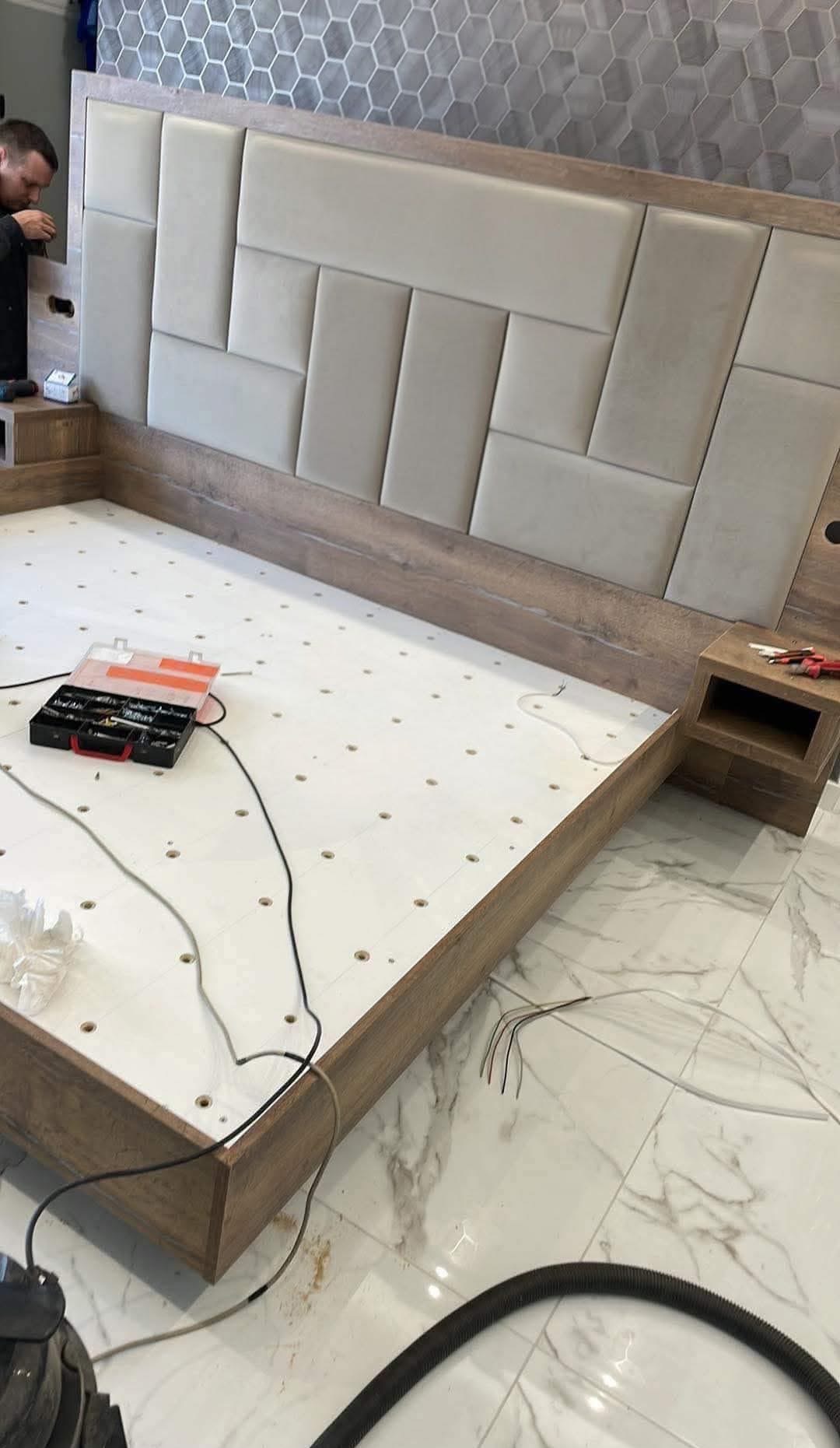 Montaż drewnianego łóżka z tapicerowanym zagłówkiem w odcieniach beżu. Widoczne narzędzia i elementy konstrukcyjne. Praca w toku.