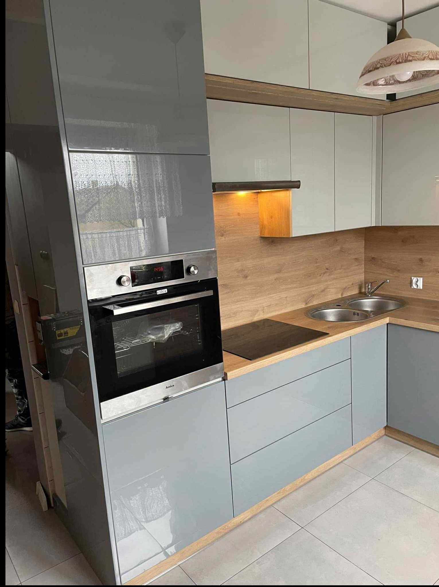 Szara kuchnia z drewnianymi akcentami: zabudowa z piekarnikiem Amica, płyta indukcyjna i zlewozmywak w blacie imitującym drewno. Nowoczesne oświetlenie.